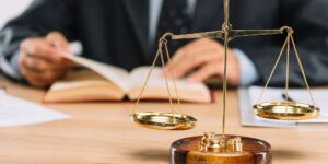 Balança em primeiro plano e no fundo, mãos de uma pessoa de terno folheia um livro em uma mesa de madeira.